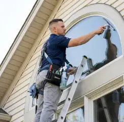 Professional window cleaner on a ladder using a squeegee to clean an exterior arched window, Detroit, MI.